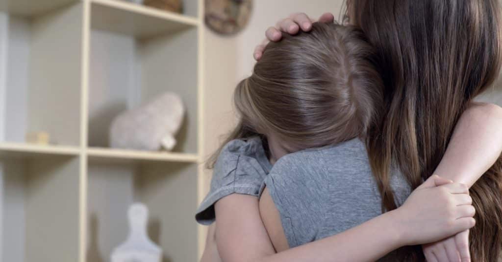 A mother hugs her upset young daughter as the daughter buries her face against her mom's shoulder in a living room.