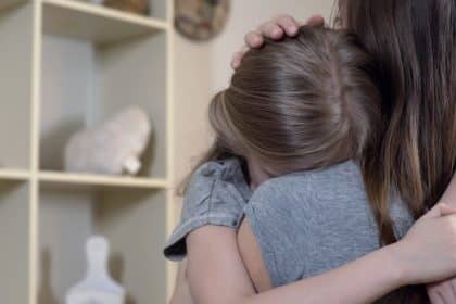 A mother hugs her upset young daughter as the daughter buries her face against her mom's shoulder in a living room.