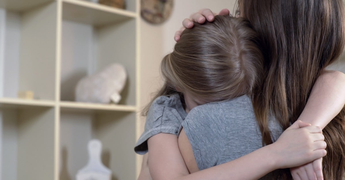 A mother hugs her upset young daughter as the daughter buries her face against her mom's shoulder in a living room.