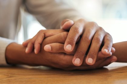 Two people gently holding hands across a wooden table, offering comfort, support, and reassurance during a conversation.