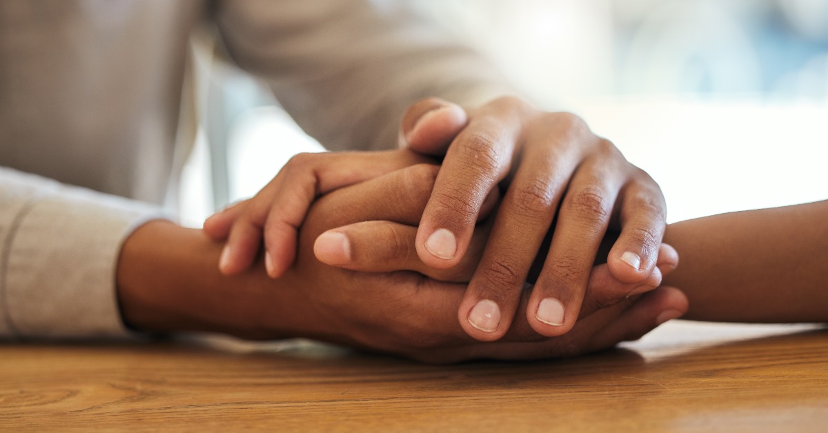 Two people gently holding hands across a wooden table, offering comfort, support, and reassurance during a conversation.