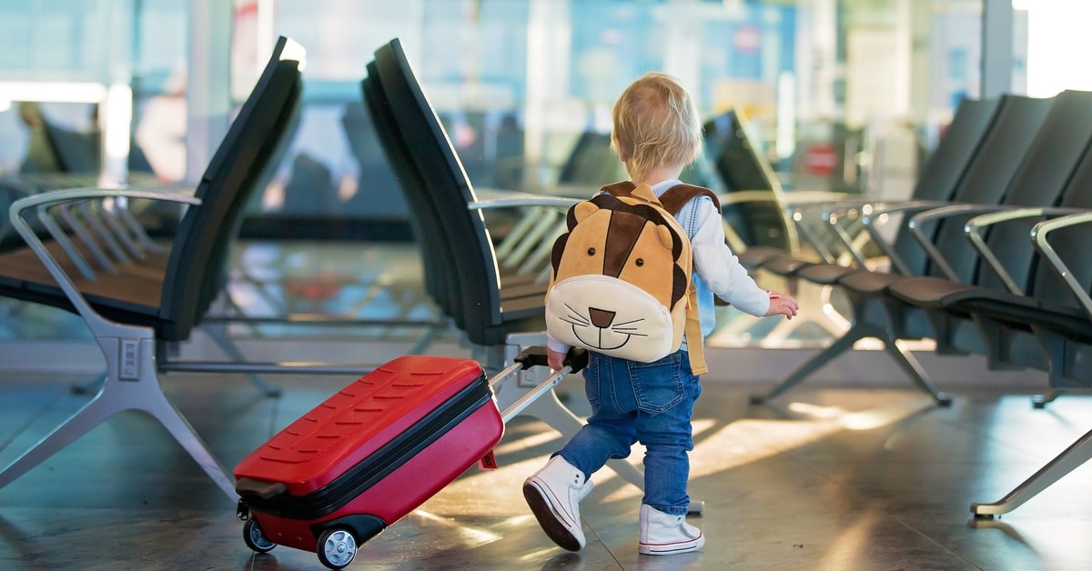 A young child wearing an animal backpack pulls a red suitcase through a bright airport terminal lined with seats.