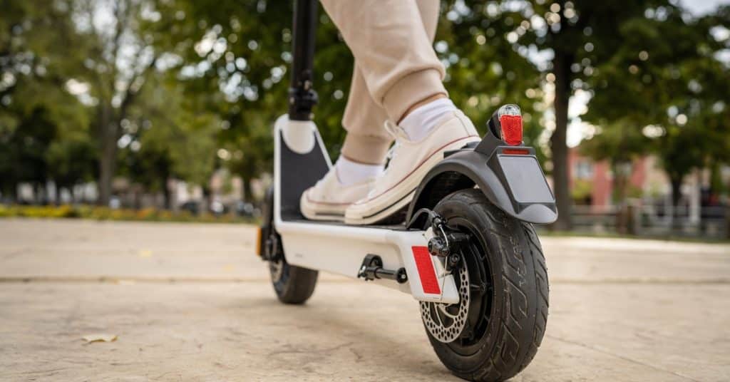A young person wearing cream-colored shoes and tan pants is standing on a white electric scooter in a park.