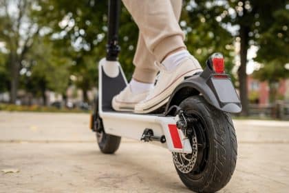 A young person wearing cream-colored shoes and tan pants is standing on a white electric scooter in a park.