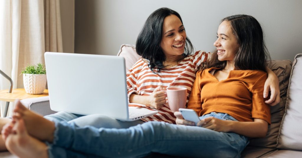 Two people smile together on a couch while using a laptop and smartphone, with one holding a pink mug.