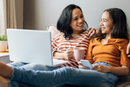 Two people smile together on a couch while using a laptop and smartphone, with one holding a pink mug.