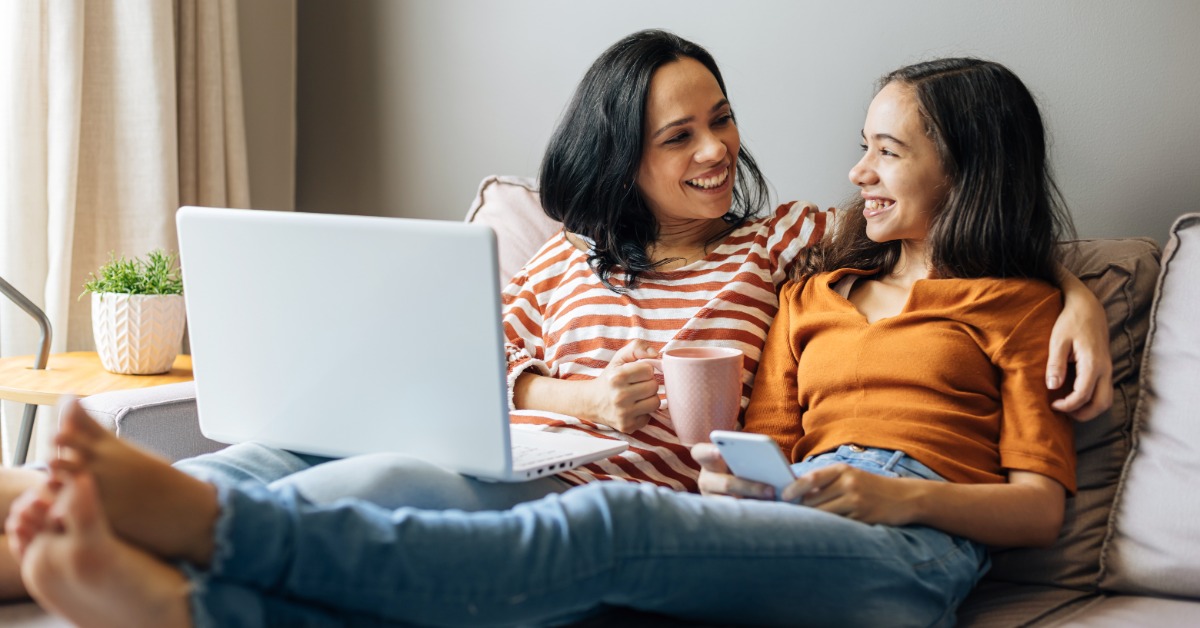 Two people smile together on a couch while using a laptop and smartphone, with one holding a pink mug.