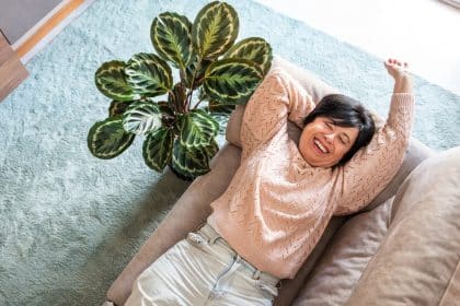 A woman is laying on a couch, smiling. There is a green plant next to her and sunlight coming in through a window.