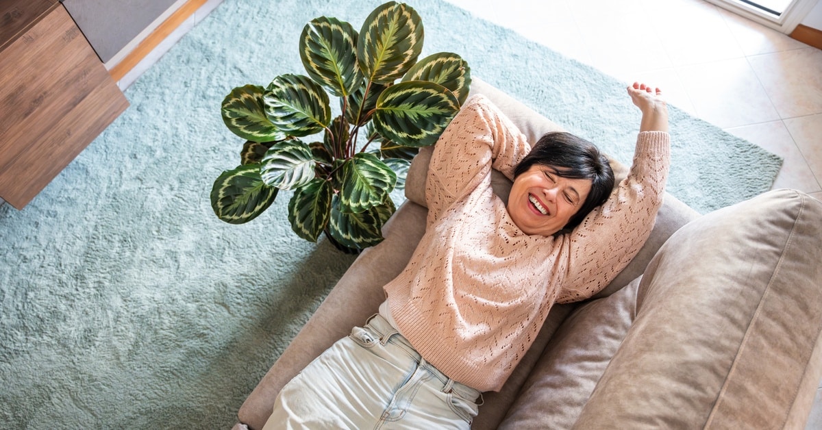 A woman is laying on a couch, smiling. There is a green plant next to her and sunlight coming in through a window.