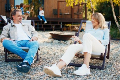 A man and woman relaxing in outdoor chairs on a gravel patio, with children and a wooden cabin behind them.