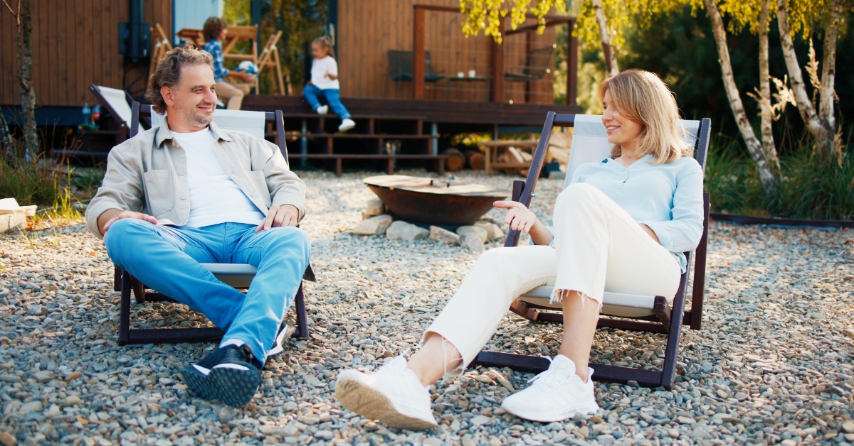 A man and woman relaxing in outdoor chairs on a gravel patio, with children and a wooden cabin behind them.