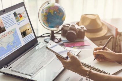 A mom sits down at the desk to plan a vacation with a notepad, computer, and her cell phone. There's a globe next to her.