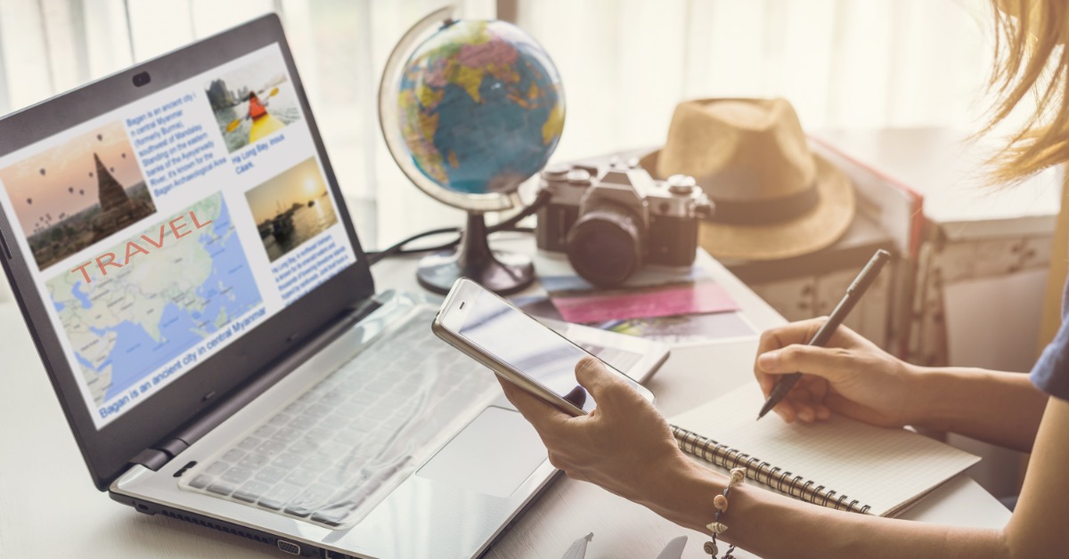 A mom sits down at the desk to plan a vacation with a notepad, computer, and her cell phone. There's a globe next to her.