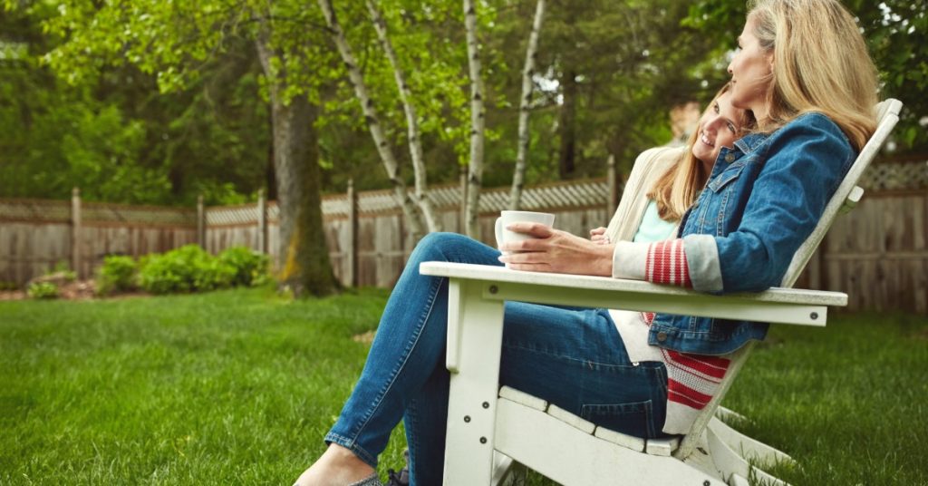 A mother and her daughter relaxing together in a white outdoor chair on a green lawn. The mother is holding a cup.