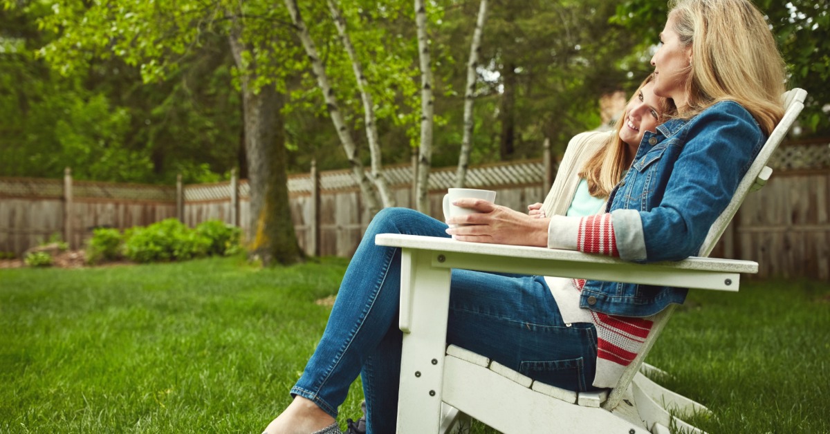 A mother and her daughter relaxing together in a white outdoor chair on a green lawn. The mother is holding a cup.