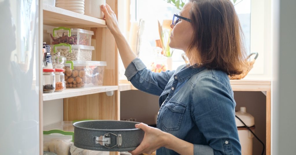 A woman in glasses reaches for a pantry canister while holding a baking pan beside shelves of food containers.