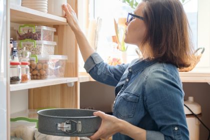 A woman in glasses reaches for a pantry canister while holding a baking pan beside shelves of food containers.