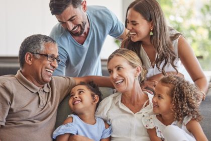 Multigenerational family of six gathers closely on a couch, smiling and laughing together in a bright living room.