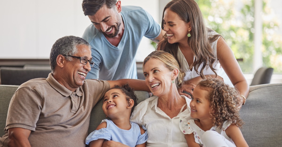Multigenerational family of six gathers closely on a couch, smiling and laughing together in a bright living room.