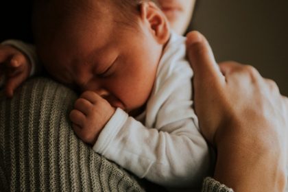  Sleeping newborn in a white outfit rests against an adult's shoulder while being gently supported by a hand.