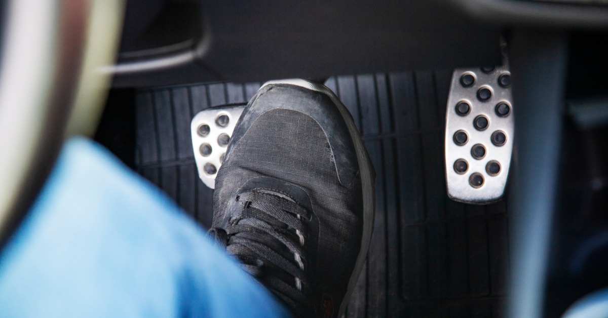 A person in the driver’s seat of a car wearing a black shoe and blue pants. The driver places a foot on the brakes.