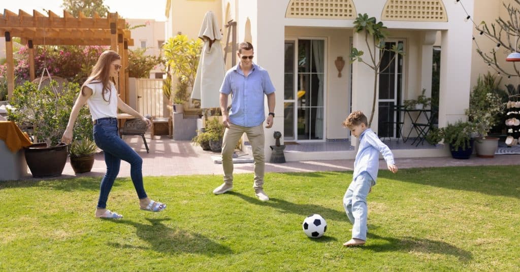 A family of three playing soccer on a grassy backyard lawn, with a young boy kicking the ball near his parents.