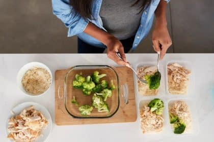 An overhead view of a woman preparing meals in containers on a counter with shredded chicken, rice, and broccoli.
