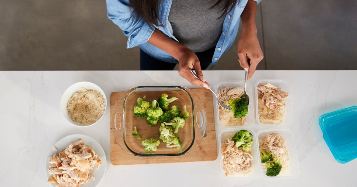 An overhead view of a woman preparing meals in containers on a counter with shredded chicken, rice, and broccoli.