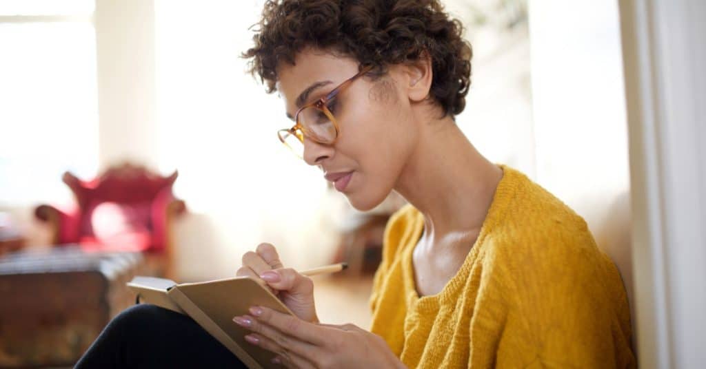A woman sits on the floor in a bright room, knees pulled toward her chest. She writes in a small notebook on her lap.