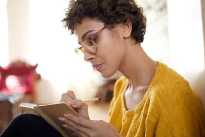 A woman sits on the floor in a bright room, knees pulled toward her chest. She writes in a small notebook on her lap.