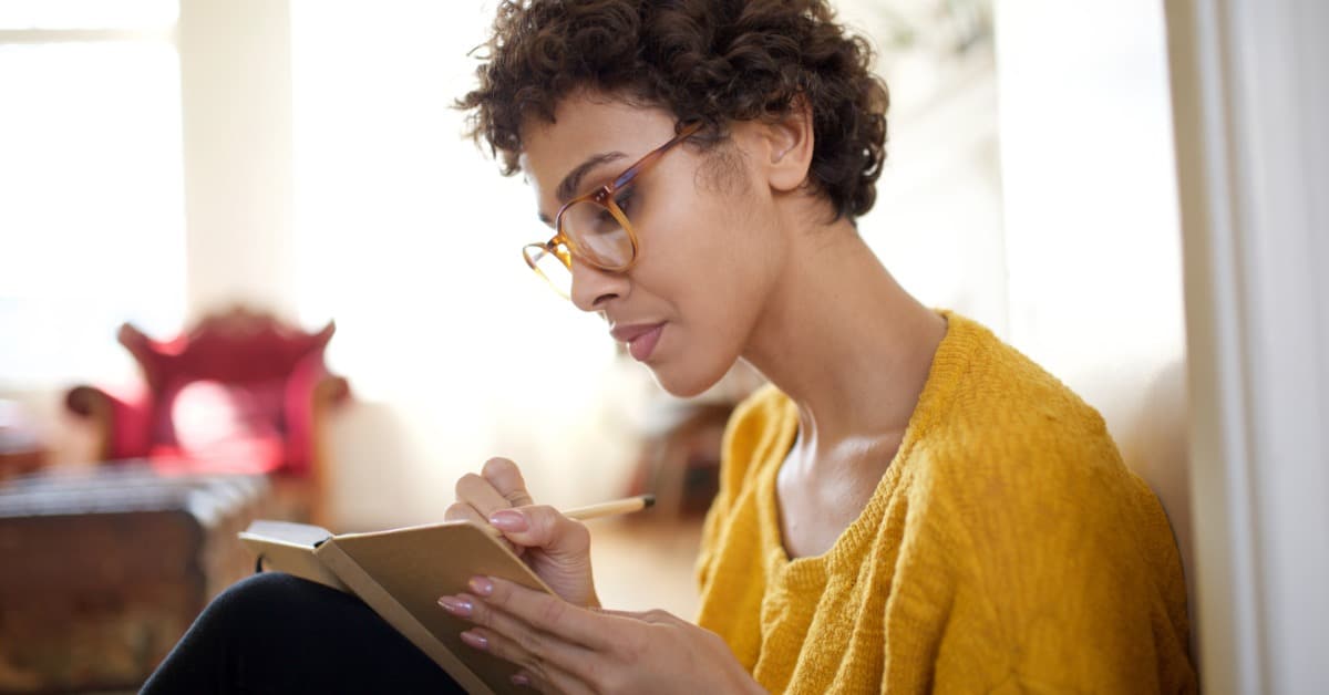 A woman sits on the floor in a bright room, knees pulled toward her chest. She writes in a small notebook on her lap.