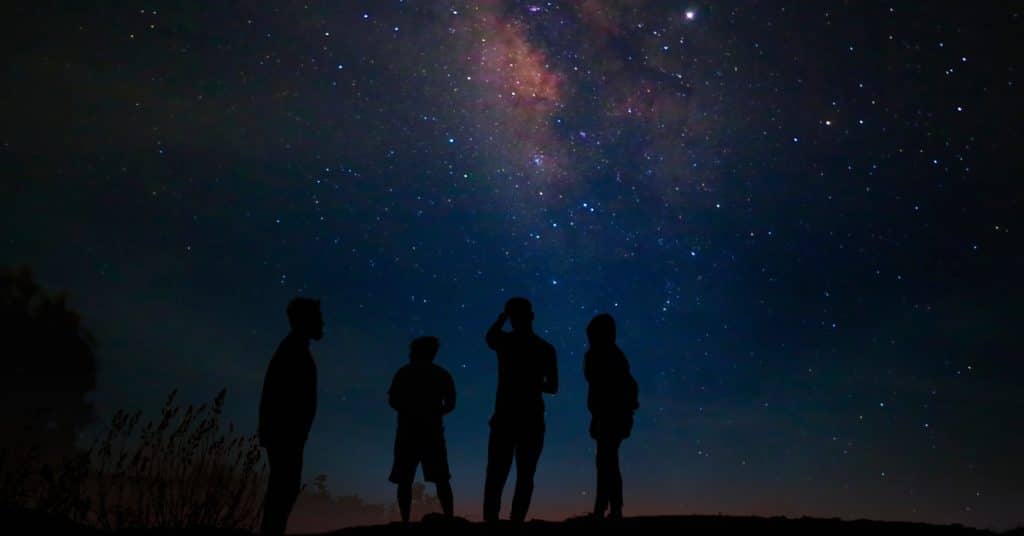 Four people stand beneath a star-filled night sky, looking toward the colorful Milky Way stretching overhead.