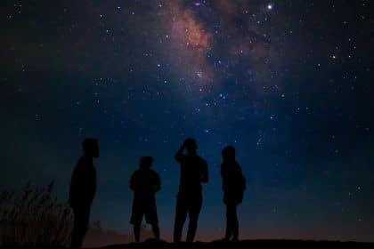 Four people stand beneath a star-filled night sky, looking toward the colorful Milky Way stretching overhead.